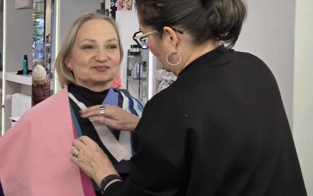 Auli tekemässä värianalyysiä – lempeä, keskittynyt ilme. Hän seisoo valoisassa tilassa ja tarkastelee värikankaita asiakkaan kasvojen läheltä.